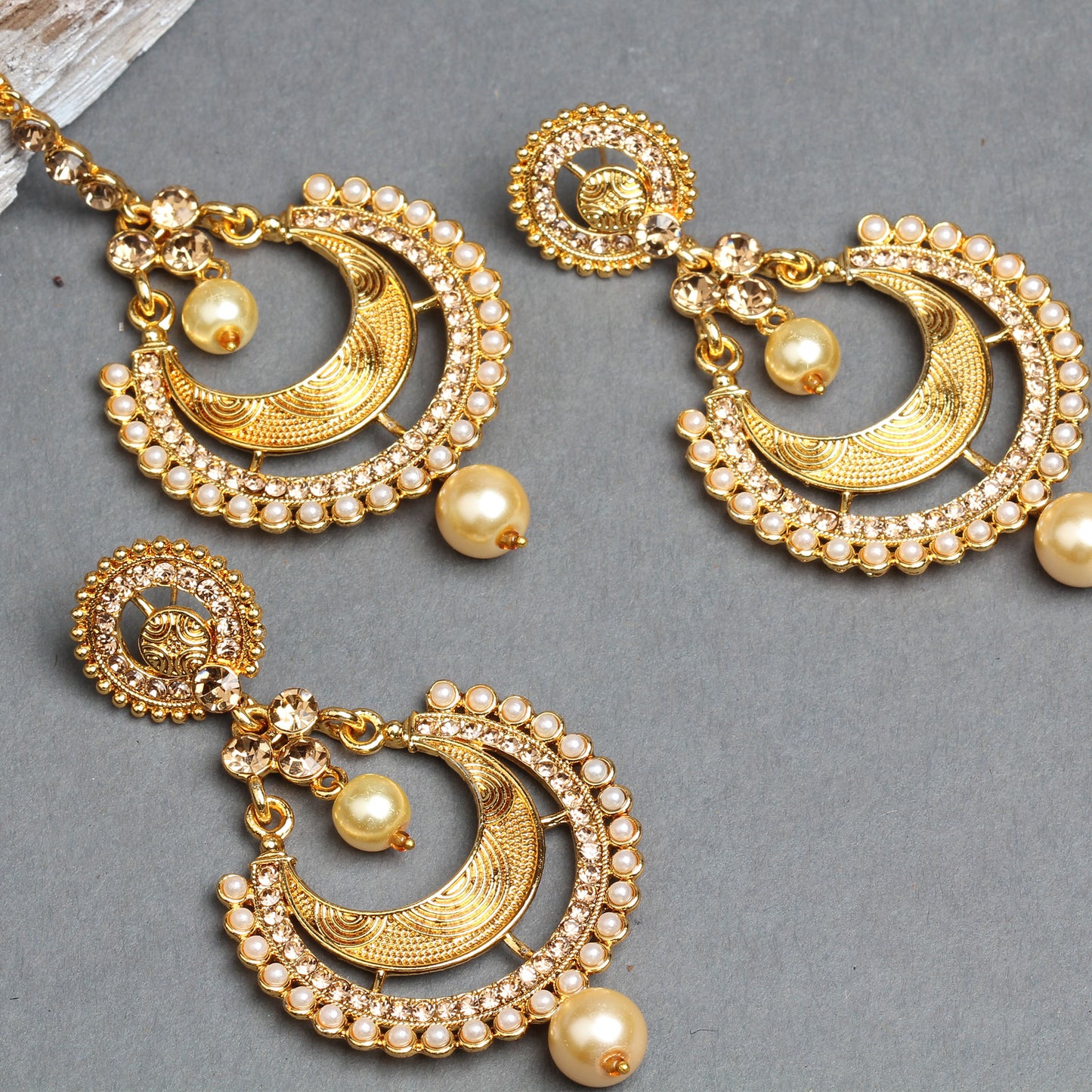 Bindhani gold-plated earrings and maang tikka set featuring crescent-shaped designs with intricate engraved detailing, sparkling white crystal stones, and a border of tiny pearl accents. The matching earrings and maang tikka are adorned with cream pearl drops and decorative crystal connectors, creating an elegant traditional bridal jewelry set. Displayed on a grey background