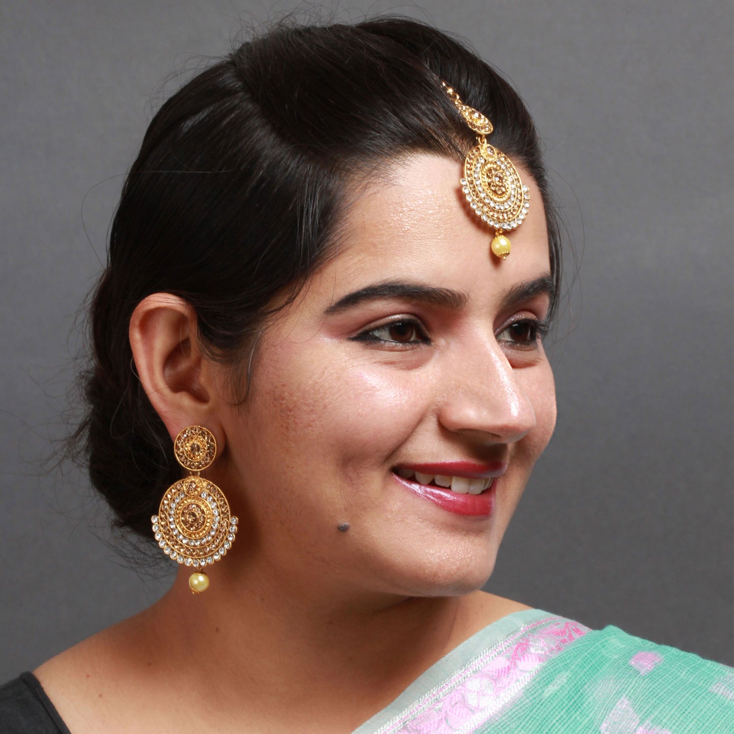 Model wearing Bindhani gold-plated statement earrings and matching maang tikka featuring layered concentric circular designs embellished with sparkling white crystal stones and intricate gold detailing. Both the earrings and forehead ornament are finished with a single cream pearl drop, creating an elegant traditional bridal look. The jewelry is styled with neatly tied black hair against a grey background.