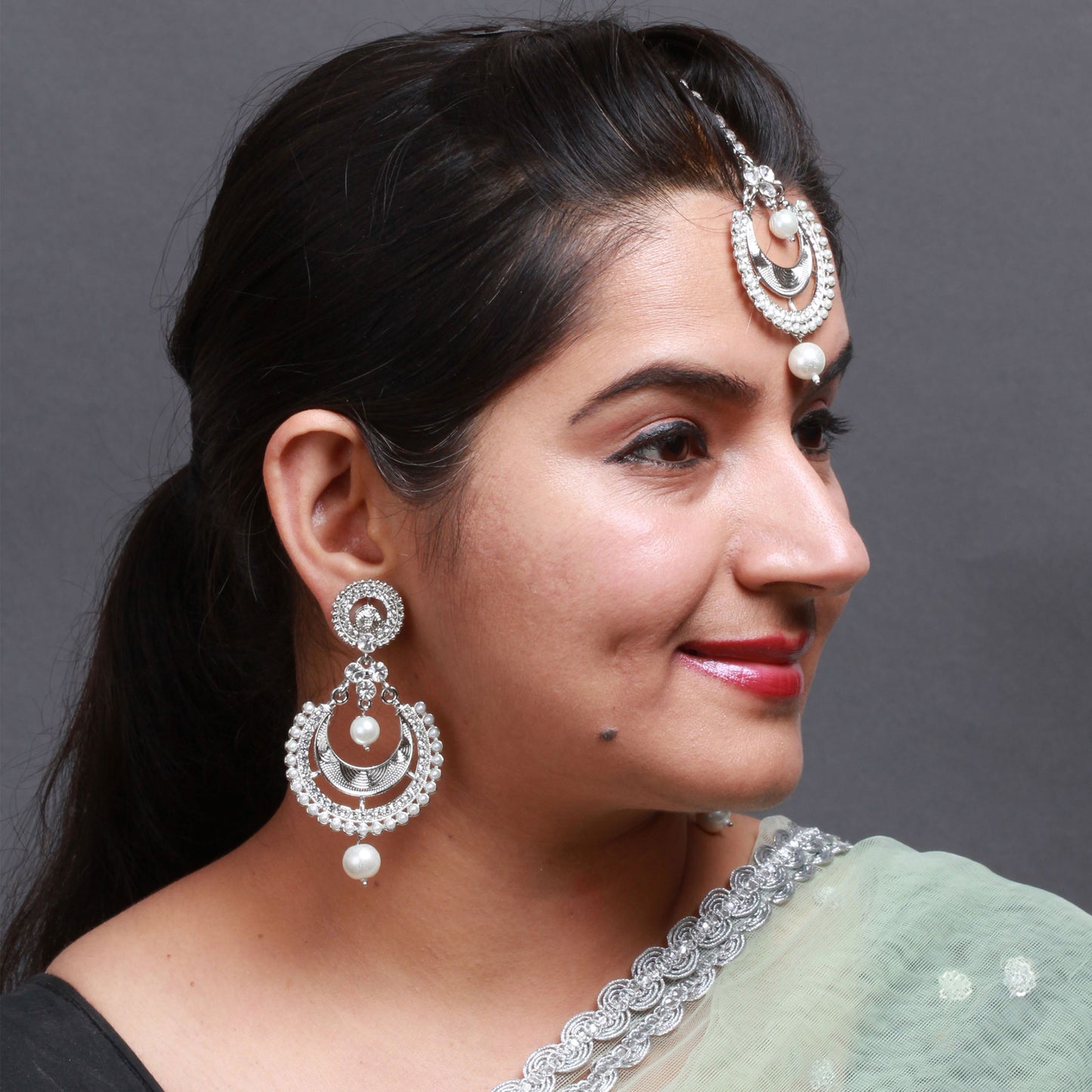 Bindhani silver-toned maang tikka and matching earrings worn on a model. The jewelry features clear stones with layered circular openwork detailing and white pearl drop accents. The maang tikka showcases a crescent-inspired layered design with a single pearl drop, while the matching earrings feature round stone-studded tops, layered circular motifs, and pearl hanging drops. Close-up view displayed against a grey background