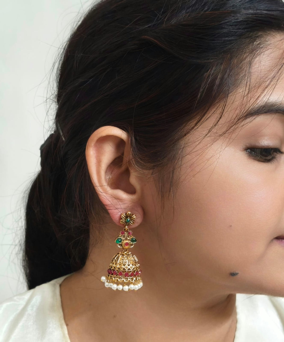 Bindhani antique gold-plated jhumka earrings featuring an intricately embossed dome design with a floral motif and a round green pink stone stud. 
The traditional temple-style earrings are adorned with engraved gold bead danglers, creating an elegant ethnic look. Lightweight drop earrings with a secure push-back closure, displayed on a white background. Perfect for weddings, festive occasions, cultural events, and traditional wear.