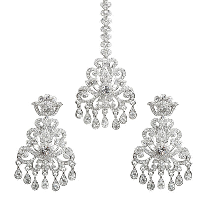 Bindhani silver plated white stone maang tikka and matching earrings set featuring an intricate floral and scrollwork design with sparkling white stones. The statement earrings showcase crown-inspired tops, detailed openwork patterns, a large central white stone, and multiple teardrop-shaped hanging white stone drops. The matching maang tikka features a decorative stone chain, ornate floral centerpiece, and dangling white stone accents. Traditional Indian bridal set displayed on a white background.