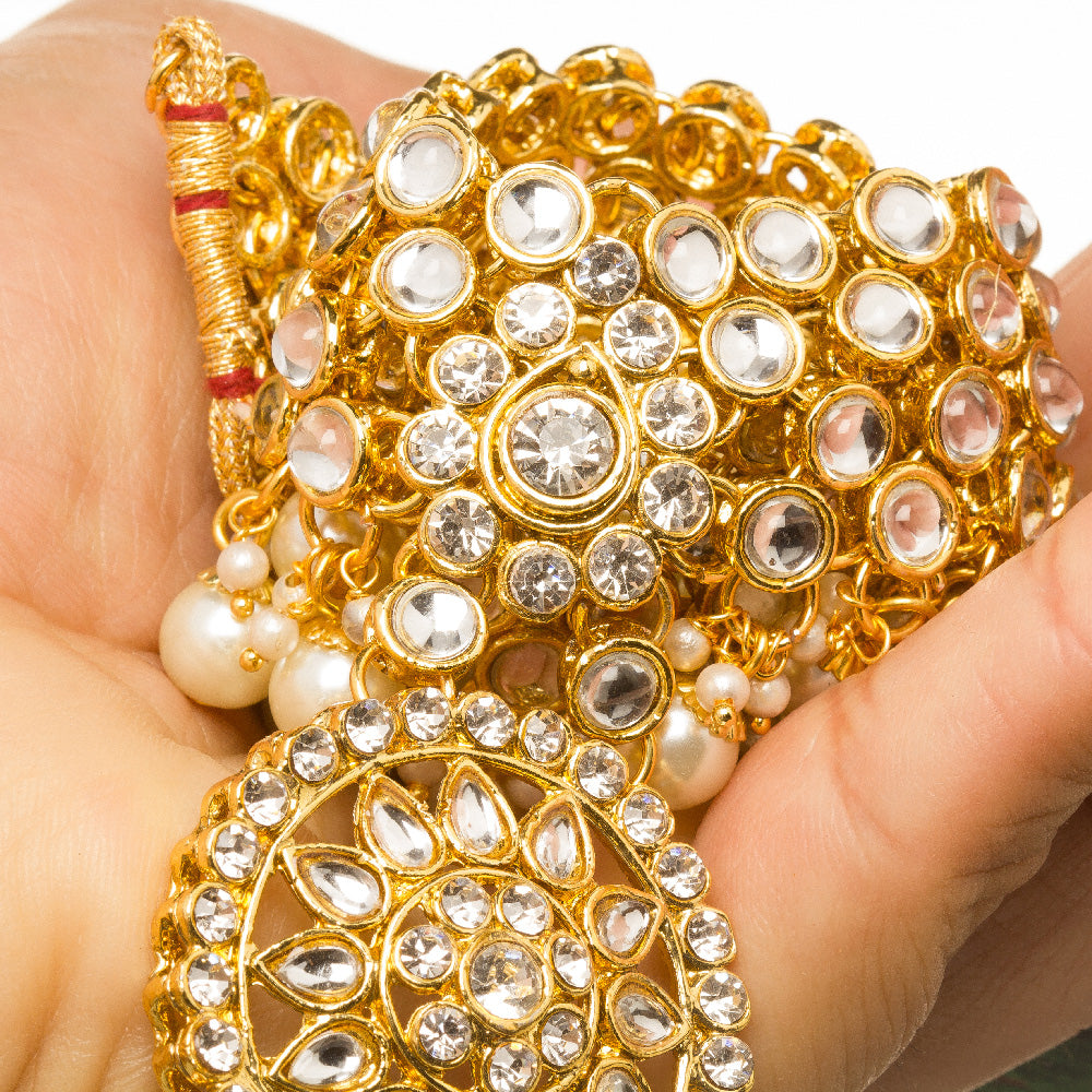 Hand holding Bindhani gold-plated kundan necklace with layered stones and pearl drops, white background"