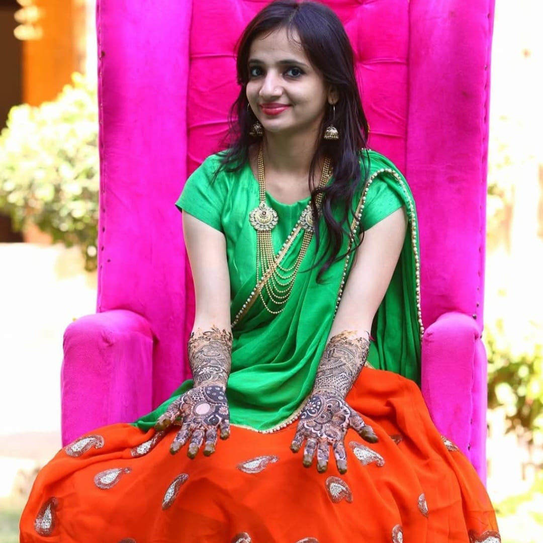 woman in green and orange traditional outfit, she is bride, sitting with smile, in her own mehandi function, teaming Bindhani ball chain haar.