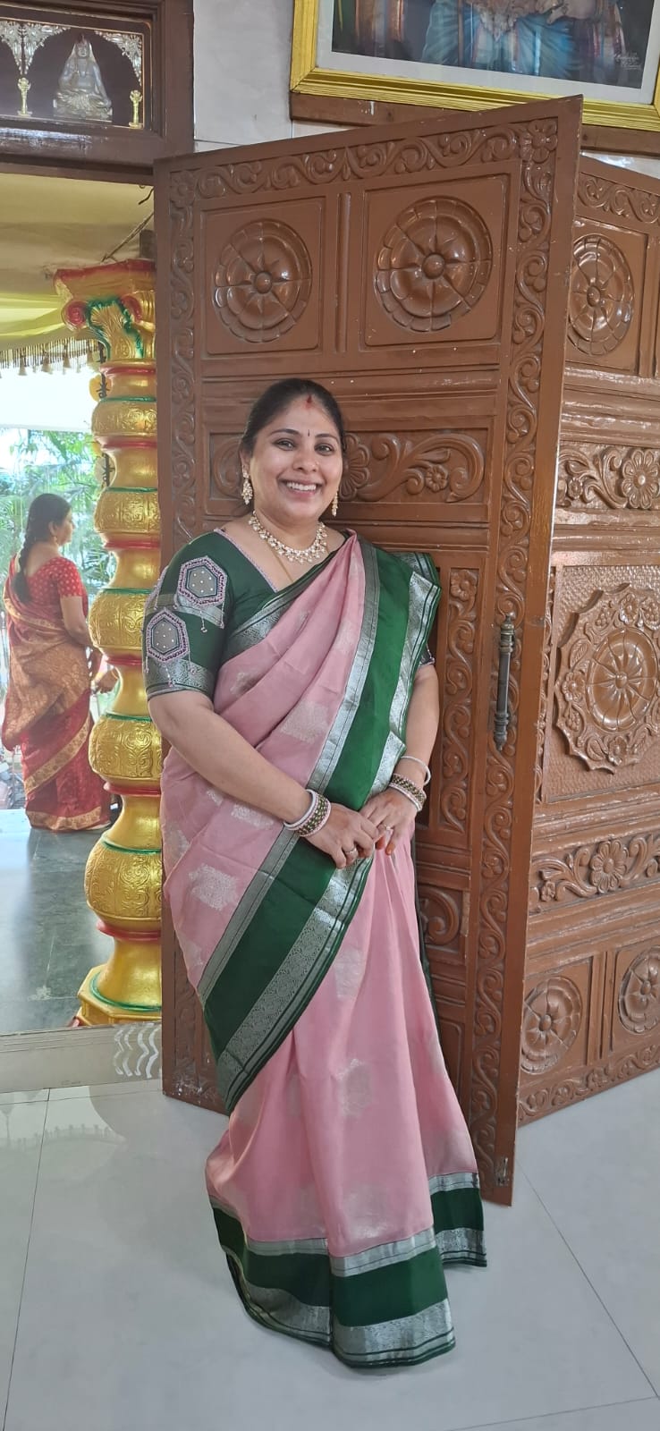 Woman in a onion  pink and green silk saree standing in front of a wooden carved door smiling and wearing Bindhani kundan necklace set.