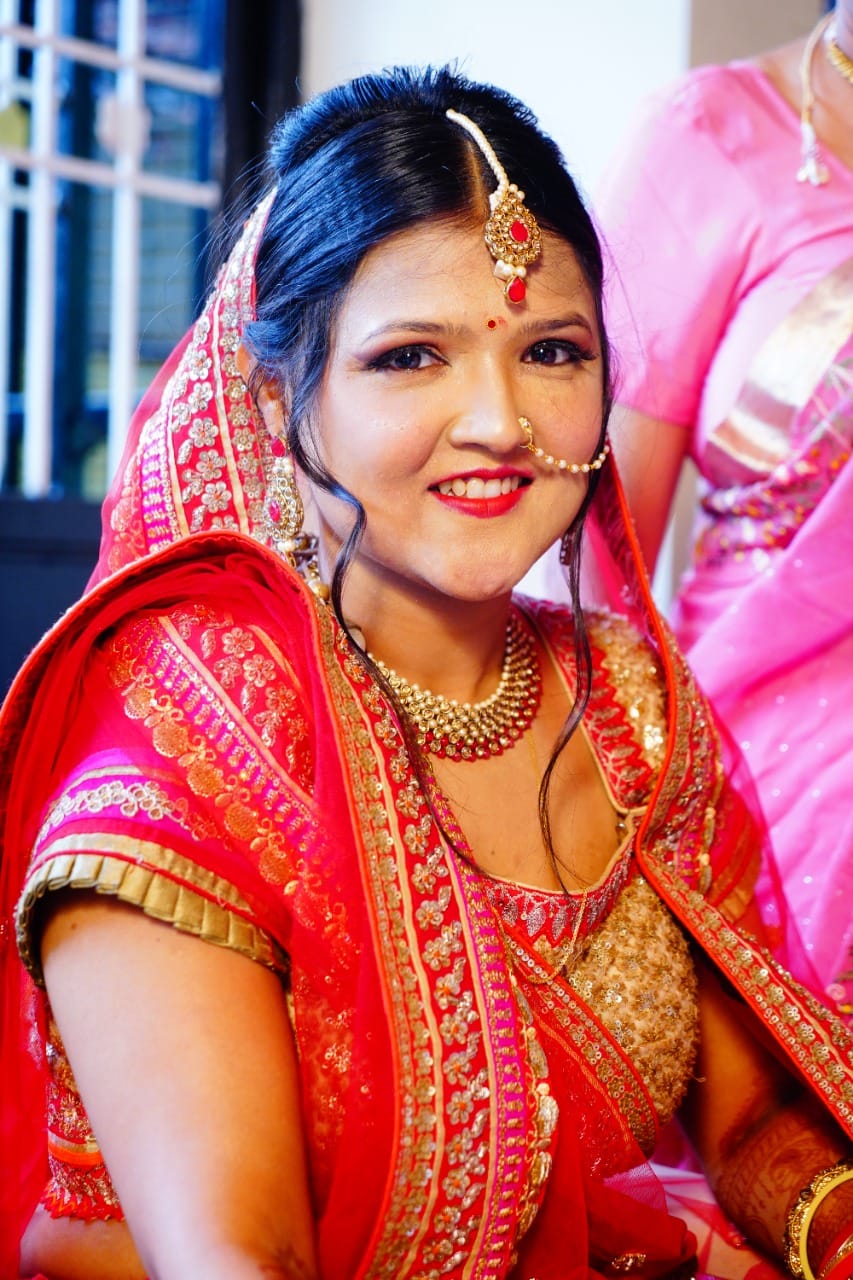 A smiling woman wearing traditional Indian bridal attire in a bright red and pink saree with gold embroidery, along with ornate gold jewelry including a maang tikka, nose ring, and layered necklaces. Nose ring and necklace set customized by Bindhani.