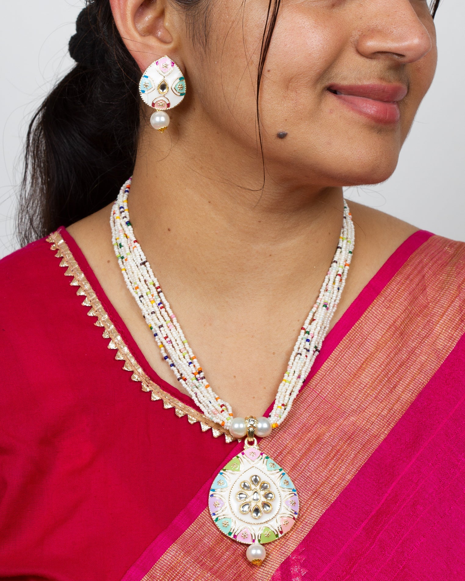 Woman wearing a Bindhani multicolor beaded necklace with a white kundan stone pendant, paired with matching enamel and pearl drop earrings, styled with a traditional pink saree.