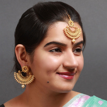 Model wearing Bindhani gold-plated earrings and matching maang tikka featuring intricate crescent-shaped designs with floral stud tops, detailed gold filigree work, and sparkling stone embellishments. The traditional jewelry set is finished with elegant pearl drops on the earrings and tikka, creating a graceful bridal look. Styled with neatly tied black hair against a grey background.
