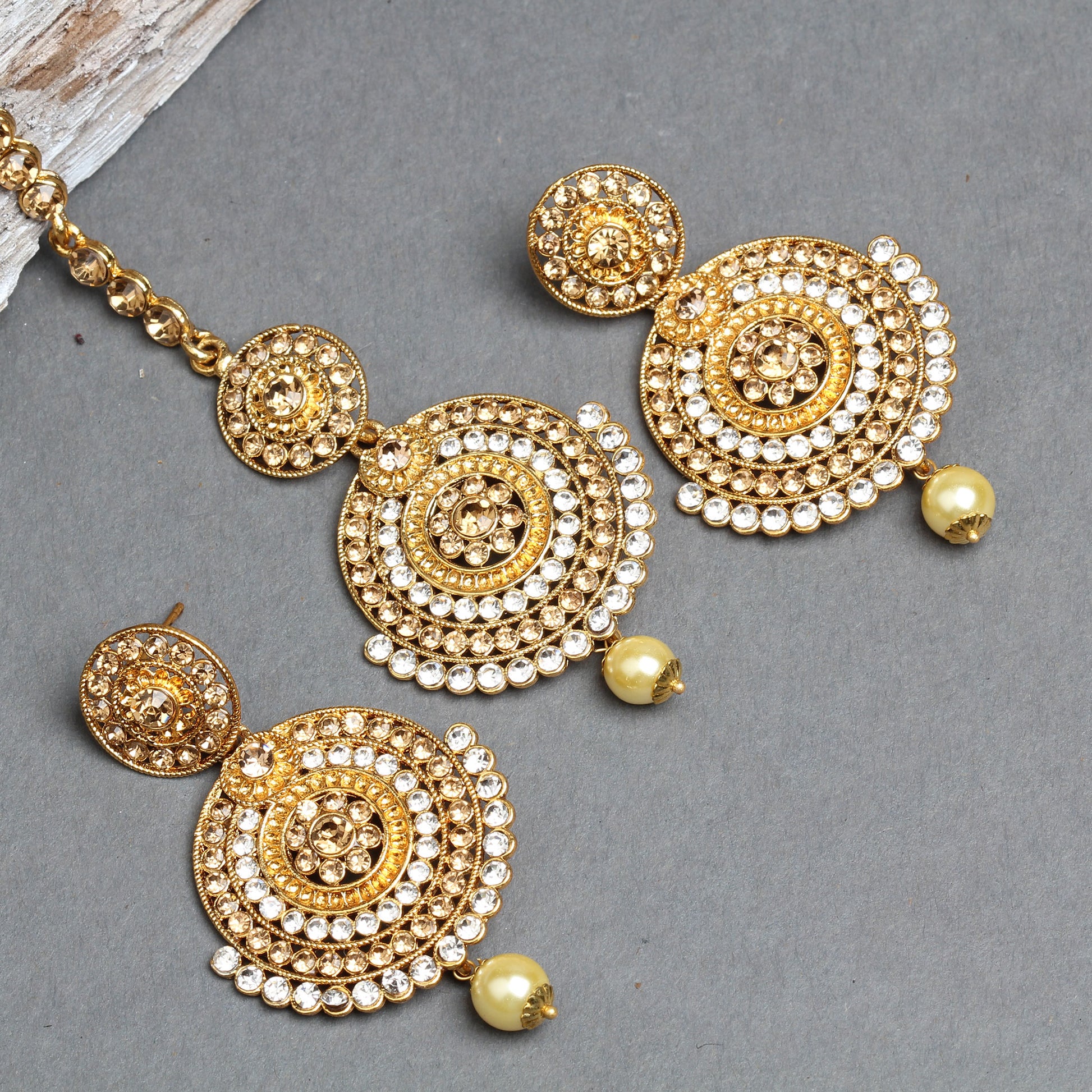 Bindhani gold-plated earrings and maang tikka set featuring layered concentric circular designs embellished with sparkling white crystal stones and intricate textured gold detailing. The matching earrings and tikka are finished with a single cream pearl drop, creating an elegant traditional bridal jewelry set. Displayed on a grey background with the stone-studded tikka chain visible.