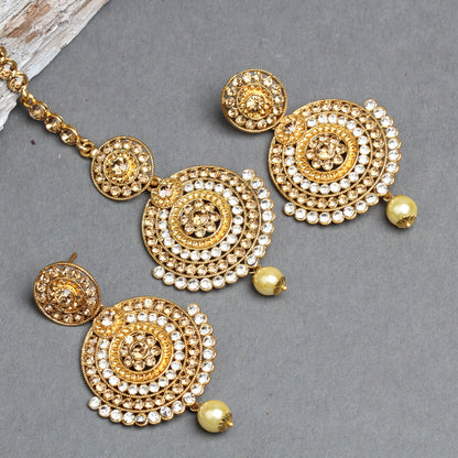 Bindhani gold-plated earrings and maang tikka set featuring layered concentric circular designs embellished with sparkling white crystal stones and intricate textured gold detailing. The matching earrings and tikka are finished with a single cream pearl drop, creating an elegant traditional bridal jewelry set. Displayed on a grey background with the stone-studded tikka chain visible.