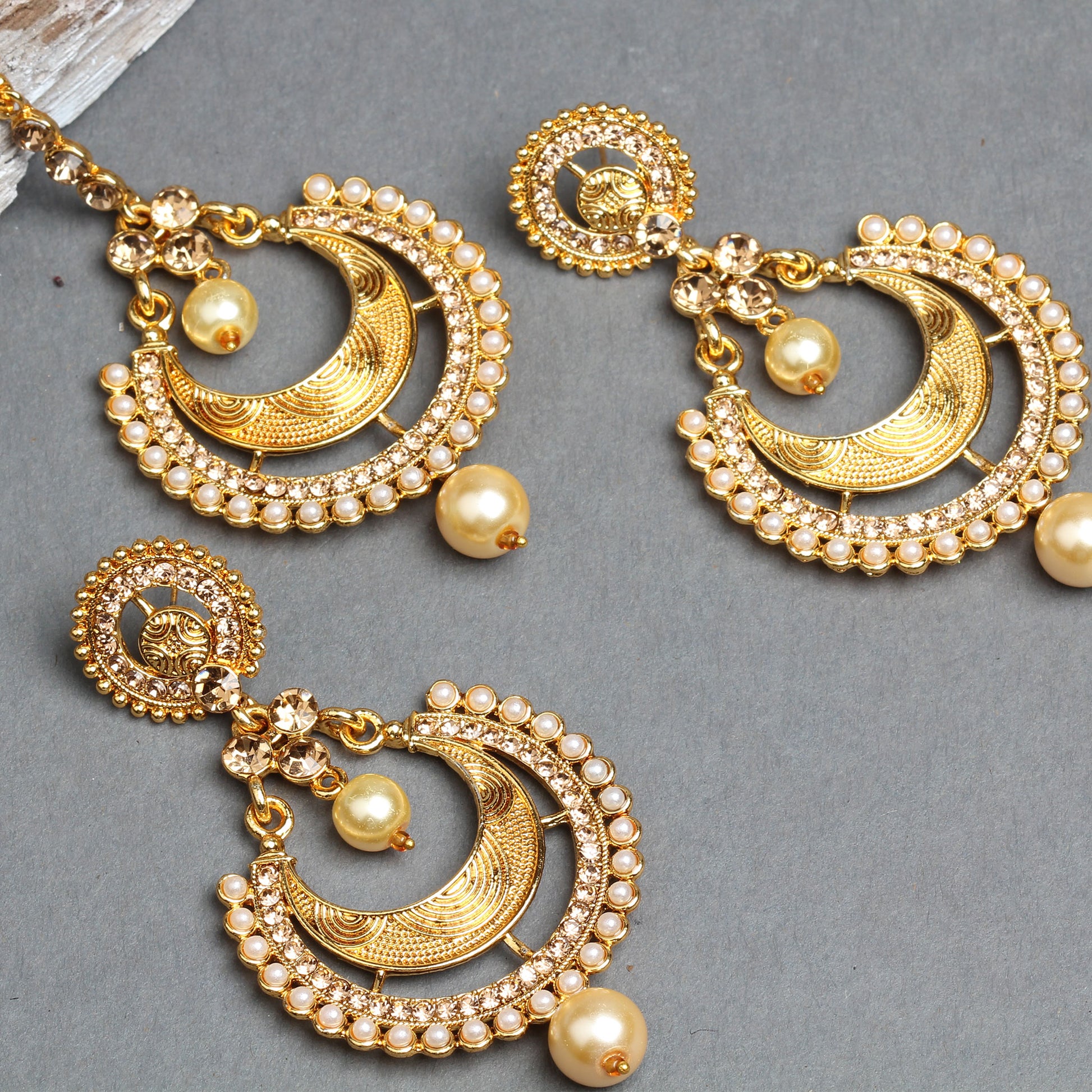 Bindhani gold-plated earrings and maang tikka set featuring crescent-shaped designs with intricate engraved detailing, sparkling white crystal stones, and a border of tiny pearl accents. The matching earrings and maang tikka are adorned with cream pearl drops and decorative crystal connectors, creating an elegant traditional bridal jewelry set. Displayed on a grey background