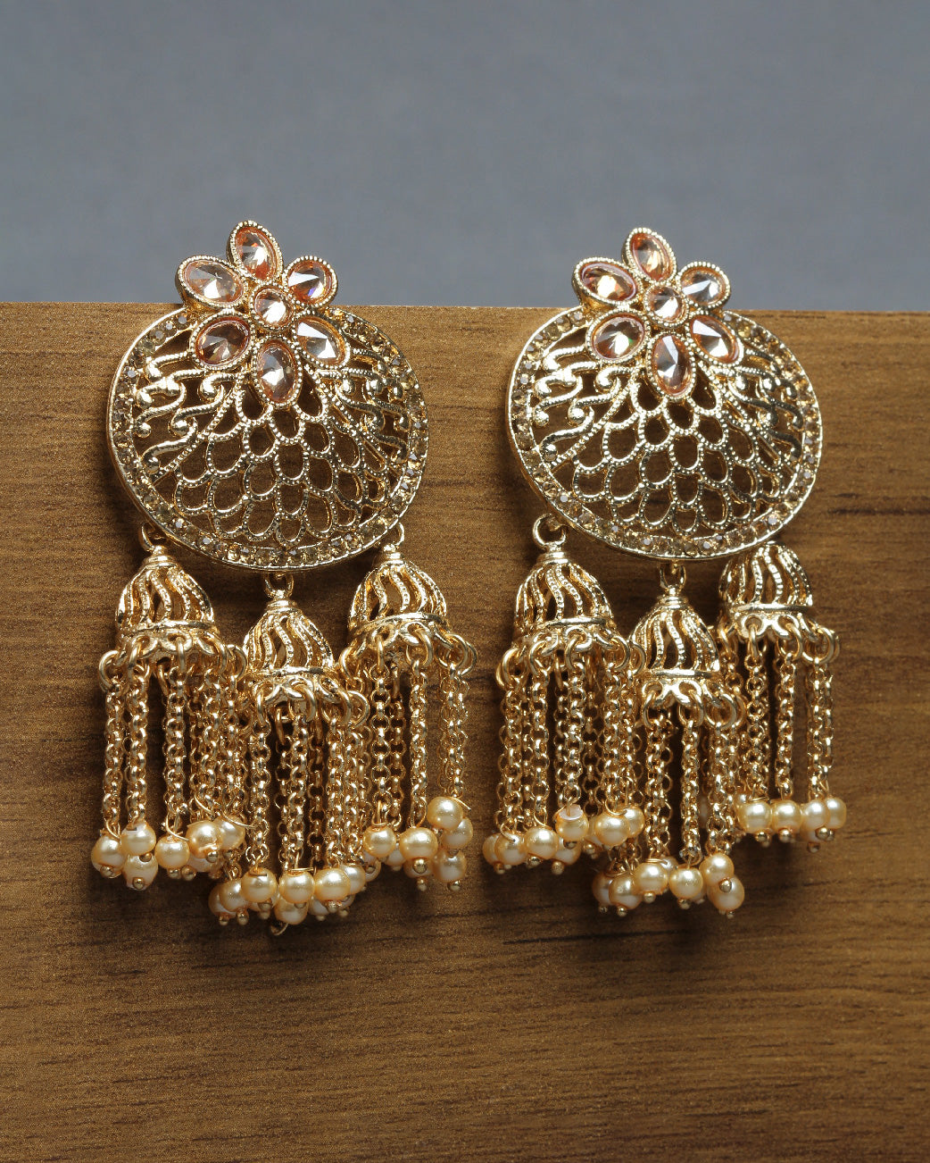 Bindhani gold-plated Jhumka earrings featuring an openwork dome design with floral motifs and teardrop-shaped stone detailing at the top. Each earring is adorned with three small jhumka tassels suspended from delicate chain strands, finished with tiny white pearl bead accents. Front view displayed on a wooden surface with a grey background.