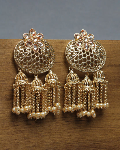 Bindhani gold-plated Jhumka earrings featuring an openwork dome design with floral motifs and teardrop-shaped stone detailing at the top. Each earring is adorned with three small jhumka tassels suspended from delicate chain strands, finished with tiny white pearl bead accents. Front view displayed on a wooden surface with a grey background.