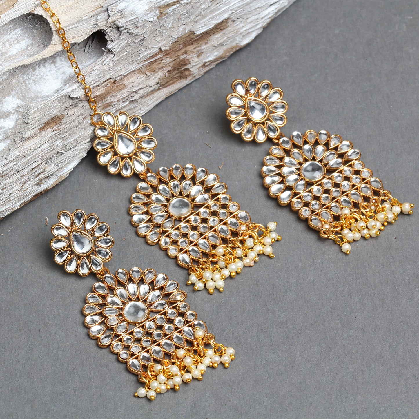 Bindhani gold-plated Kundan jewelry set featuring a floral maang tikka and matching long statement earrings. The design showcases white teardrop and oval Kundan stones arranged in floral motifs with a central oval stone, cascading Kundan detailing, and pearl bead tassels. The jewelry is displayed on a grey textured background with a decorative wooden prop.