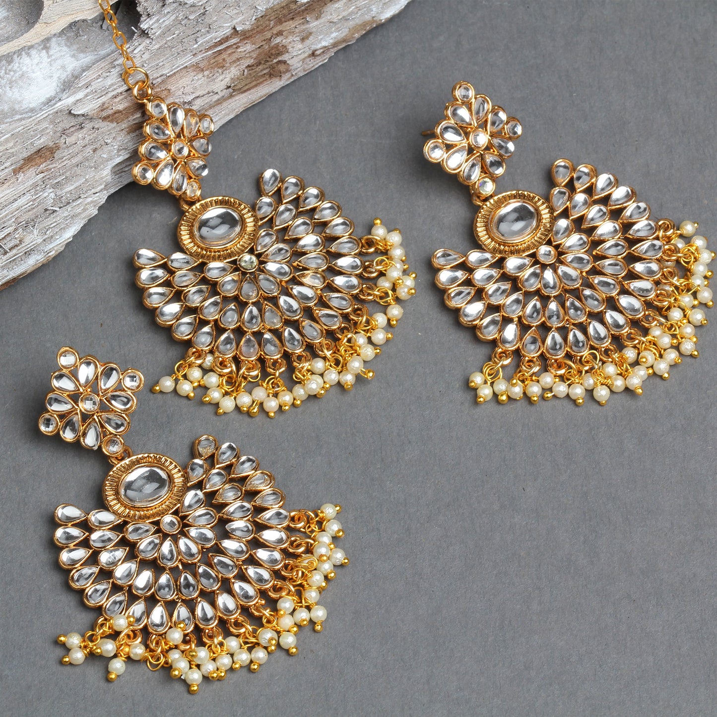 Bindhani gold-plated Kundan jewelry set featuring a floral maang tikka and matching chandbali earrings. The earrings showcase white teardrop Kundan stones arranged in a fan-shaped floral design with a central oval stone, intricate gold detailing, and pearl bead tassels. The matching maang tikka mirrors the same design with pearl bead fringes. Flat lay displayed on a grey textured background with a decorative wooden prop.
