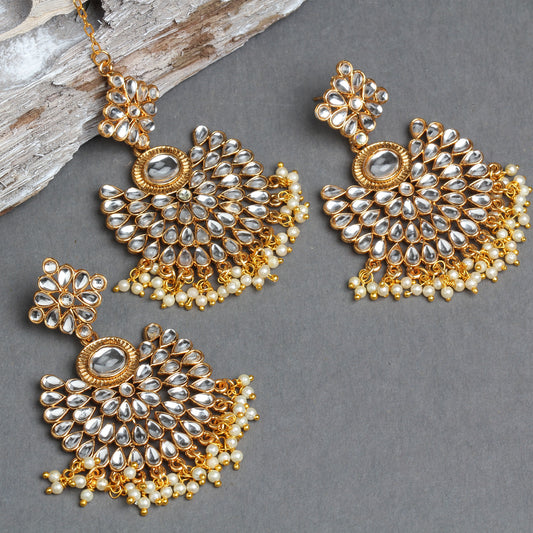 Bindhani gold-plated Kundan jewelry set featuring a floral maang tikka and matching chandbali earrings. The earrings showcase white teardrop Kundan stones arranged in a fan-shaped floral design with a central oval stone, intricate gold detailing, and pearl bead tassels. The matching maang tikka mirrors the same design with pearl bead fringes. Flat lay displayed on a grey textured background with a decorative wooden prop.