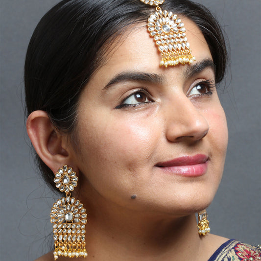 Female model wearing a Bindhani gold-plated Kundan maang tikka and matching statement earrings. The maang tikka features a floral top with white Kundan stones, cascading rows of Kundan detailing, and pearl bead drops. The long earrings have floral studs with layered Kundan motifs and pearl bead fringes. Close-up side profile showcasing the jewelry against a grey background.