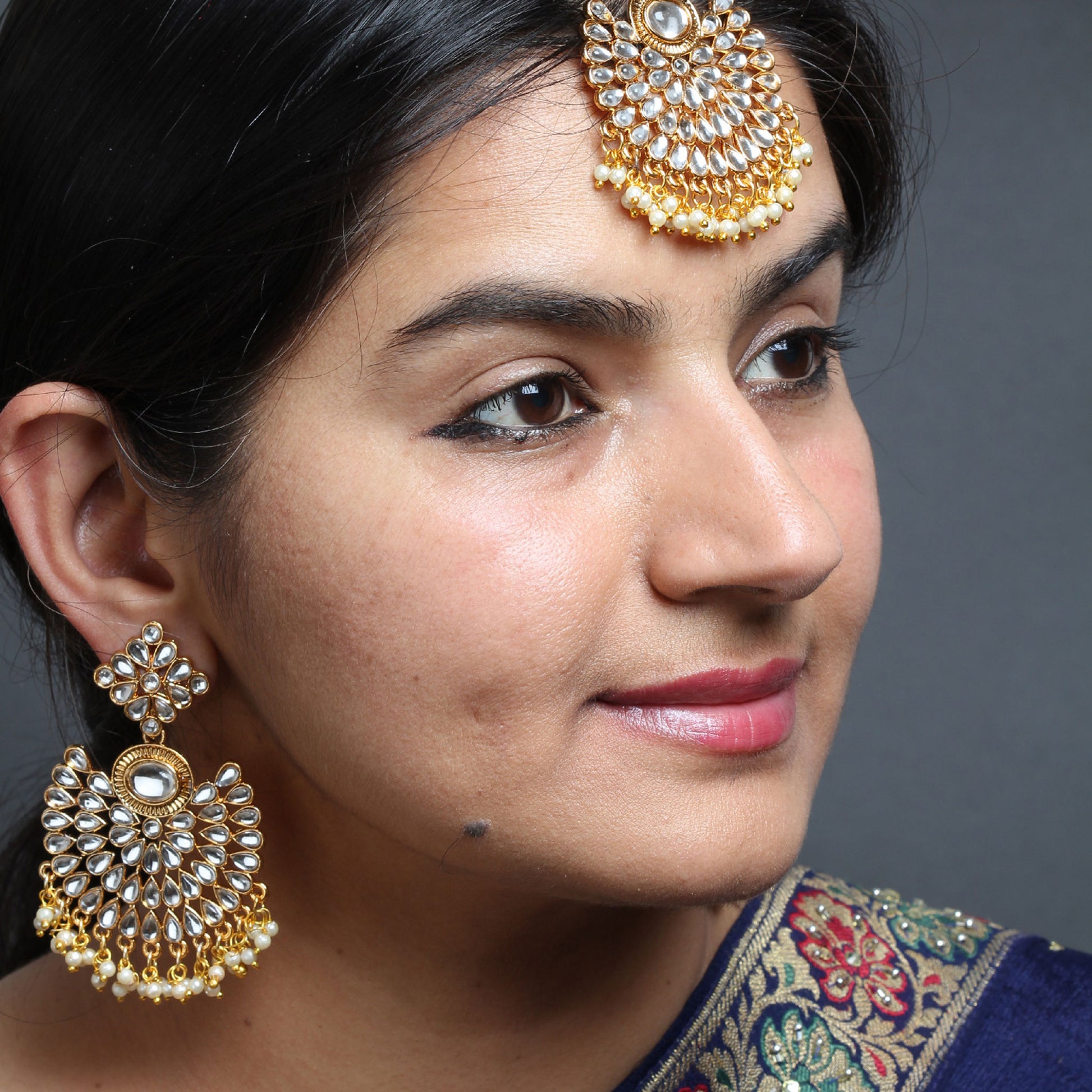 Close-up of a female model wearing a Bindhani gold-plated Kundan maang tikka and matching statement earrings. The maang tikka features concentric floral Kundan motifs with pearl bead fringes. The large round chandbali-style earrings showcase white teardrop Kundan stones, a central oval stone, intricate gold detailing, and pearl bead tassels. Side profile view against a grey background.