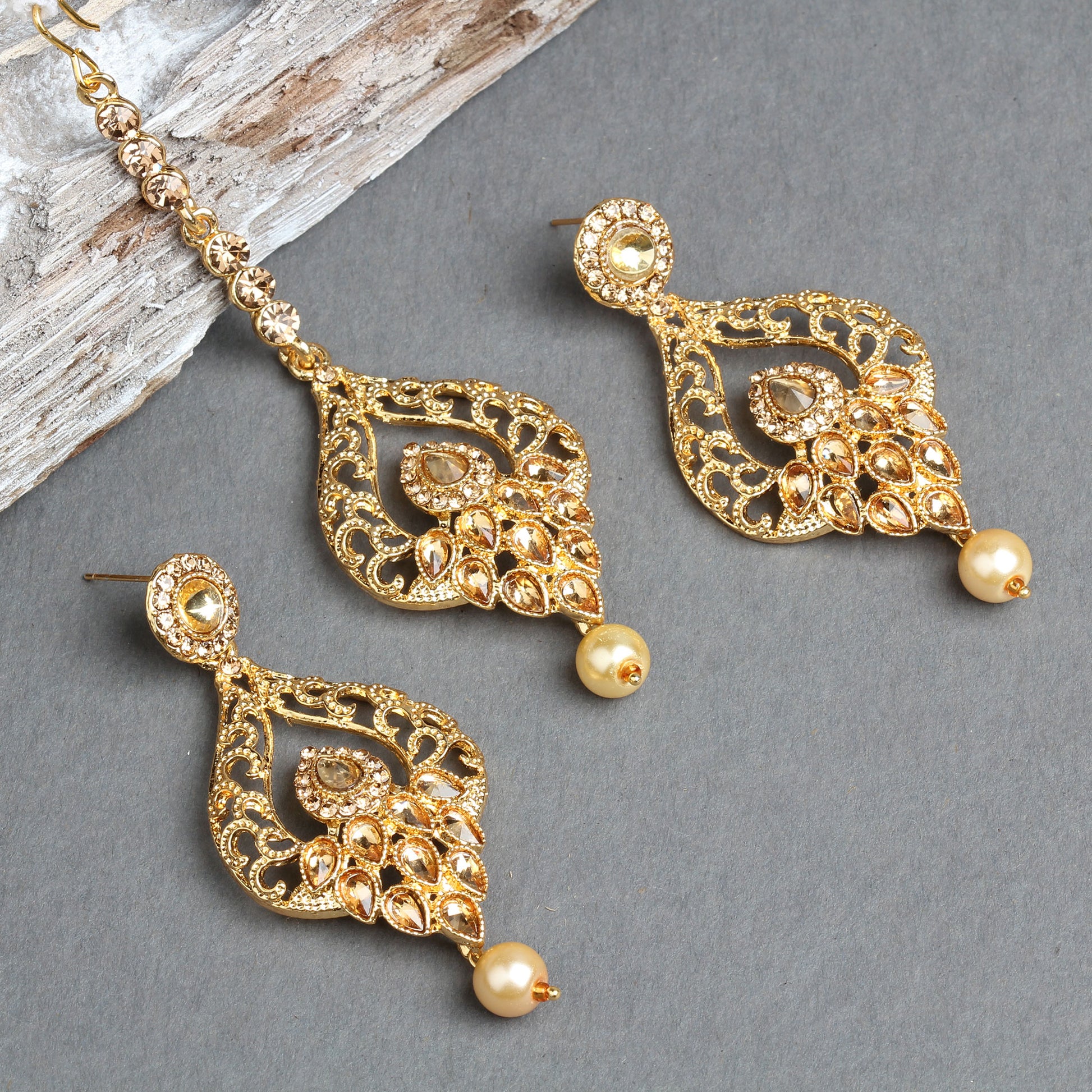 Bindhani gold-plated earrings and maang tikka set featuring leaf-shaped openwork designs with intricate filigree detailing, floral motifs, and sparkling crystal stone embellishments. The matching earrings and tikka are finished with elegant cream pearl drops, creating a traditional bridal jewelry look. Displayed on a grey background with a crystal-studded maang tikka chain visible.
