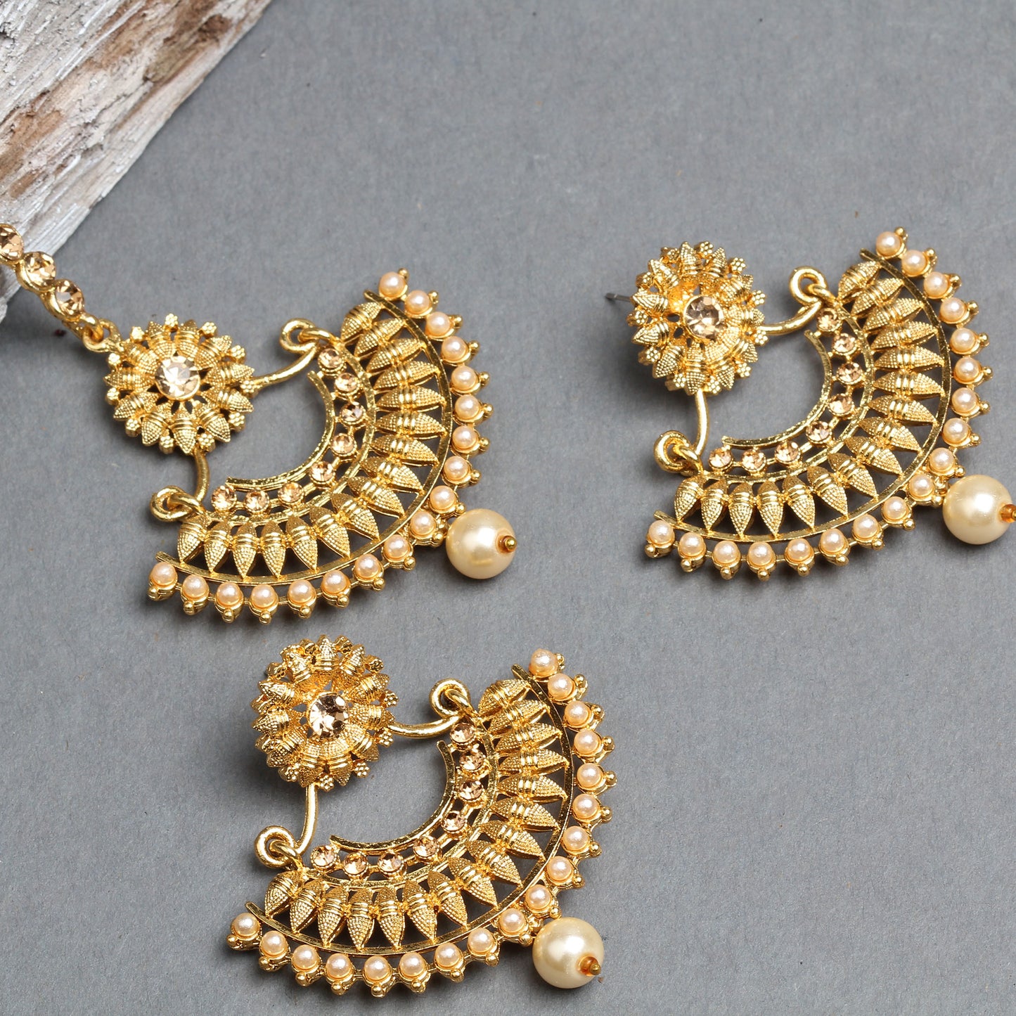 Bindhani gold-plated earrings and maang tikka set featuring elegant crescent-shaped designs with floral stud tops, intricate gold filigree detailing, and sparkling stone embellishments. The jewelry is decorated with a border of small cream pearl beads and finished with a single large pearl drop, creating a traditional bridal look. Displayed on a grey background.