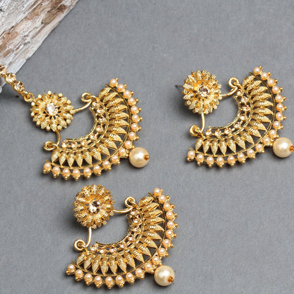 Bindhani gold-plated earrings and maang tikka set featuring elegant crescent-shaped designs with floral stud tops, intricate gold filigree detailing, and sparkling stone embellishments. The jewelry is decorated with a border of small cream pearl beads and finished with a single large pearl drop, creating a traditional bridal look. Displayed on a grey background.