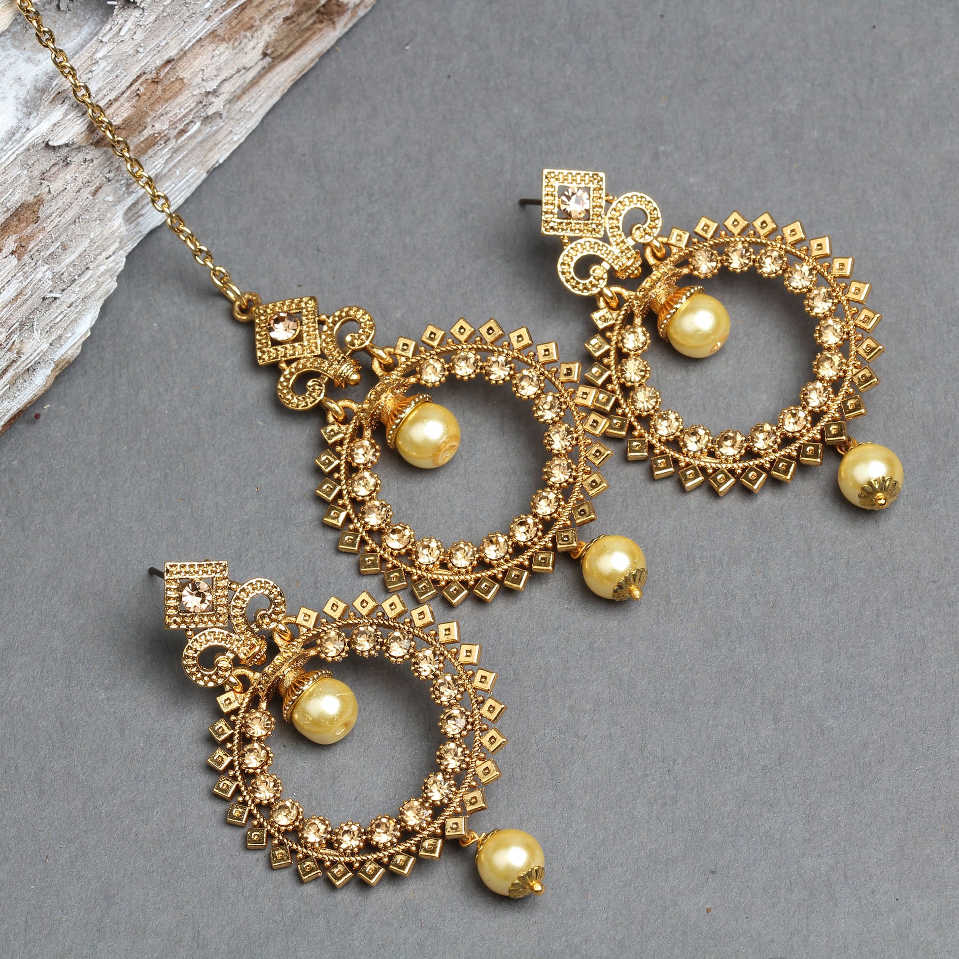 Bindhani gold-plated earrings and maang tikka set featuring circular open-hoop designs embellished with sparkling white crystal stones and geometric textured detailing. The matching earrings and maang tikka are accented with decorative square and scroll motifs and finished with two cream pearl drops on each piece, creating an elegant traditional bridal jewelry set. Displayed on a grey background with the maang tikka chain  S Hook visible