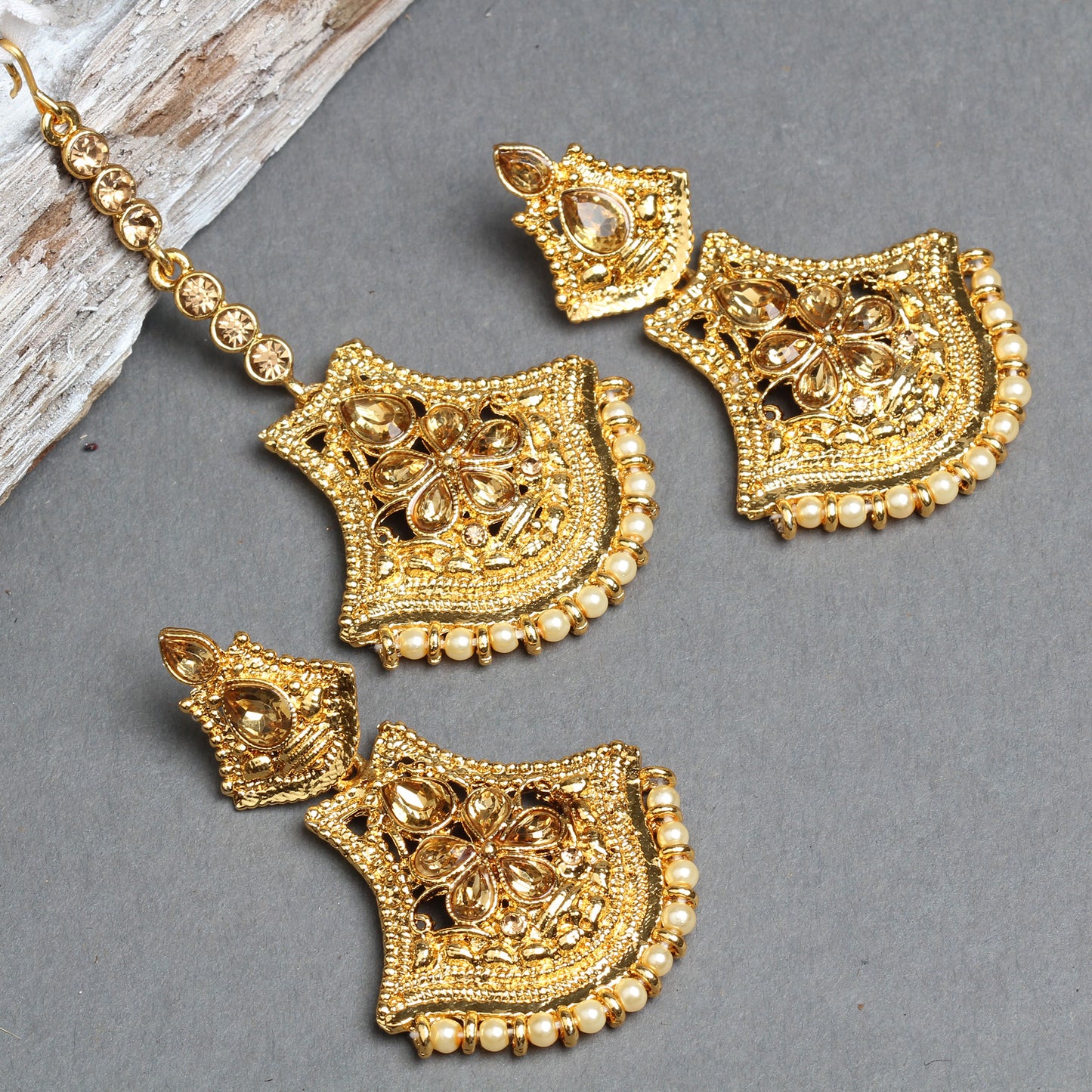 Bindhani gold-plated earrings and maang tikka set featuring shield-shaped floral designs with teardrop-shaped champagne crystal stones, intricate openwork detailing, and textured gold craftsmanship. The matching earrings and maang tikka are finished with a border of tiny cream pearl beads, creating an elegant traditional bridal jewelry set. Displayed on a grey background
