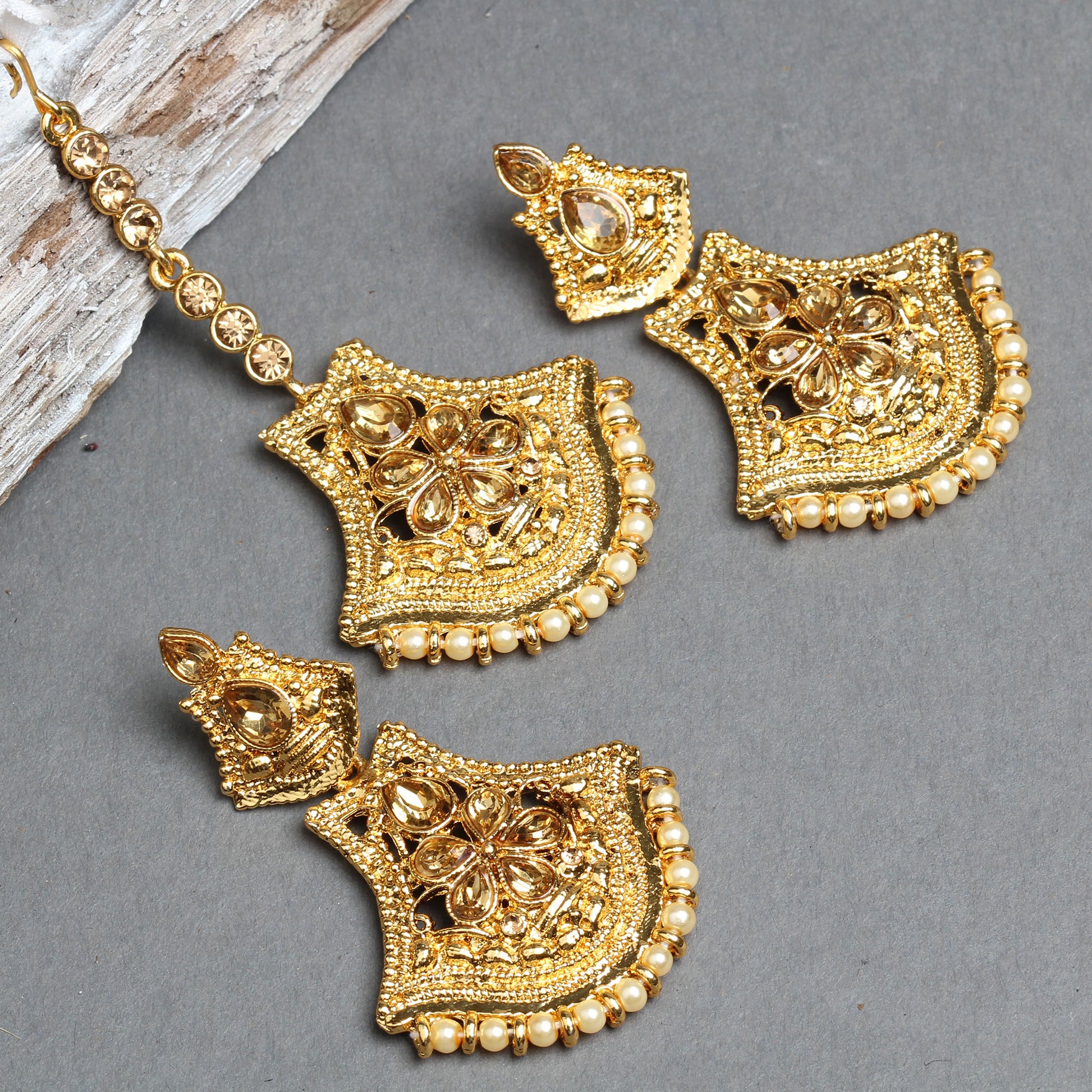 Bindhani gold-plated earrings and maang tikka set featuring shield-shaped floral designs with teardrop-shaped champagne crystal stones, intricate openwork detailing, and textured gold craftsmanship. The matching earrings and maang tikka are finished with a border of tiny cream pearl beads, creating an elegant traditional bridal jewelry set. Displayed on a grey background