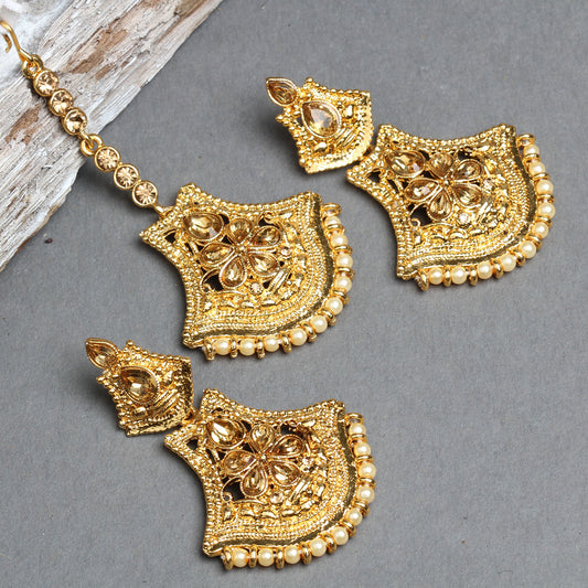 Bindhani gold-plated earrings and maang tikka set featuring shield-shaped floral designs with teardrop-shaped champagne crystal stones, intricate openwork detailing, and textured gold craftsmanship. The matching earrings and maang tikka are finished with a border of tiny cream pearl beads, creating an elegant traditional bridal jewelry set. Displayed on a grey background