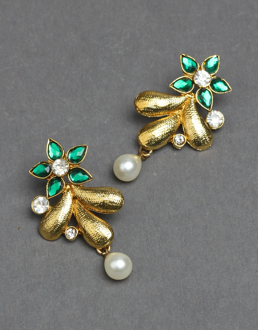 Bindhani gold-plated floral drop earrings featuring a flower design with emerald green petal-shaped stones and a central round white stone. The earrings include textured gold leaf-shaped accents, small white stone details, and elegant pearl drop hangings for a graceful traditional look. Displayed as a pair on a dark grey background.