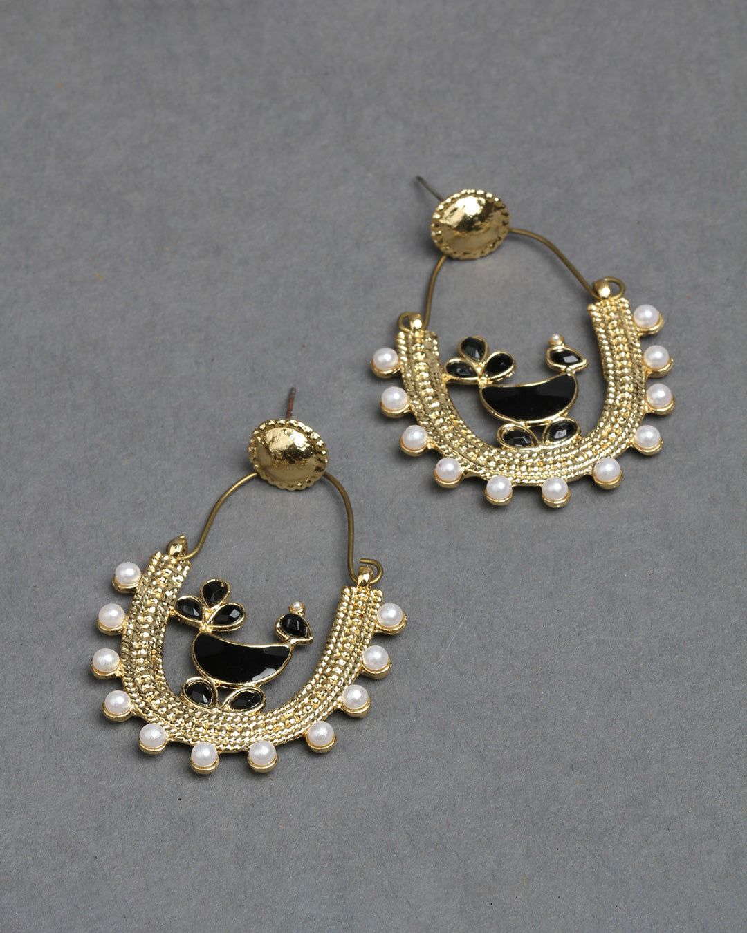 Bindhani gold-plated hoop earrings featuring a traditional peacock design with black enamel detailing at the center. The U-shaped earrings are decorated with intricate gold beadwork, white pearl drops along the outer edge, and a round textured stud top. Elegant handcrafted ethnic earrings displayed as a pair on a dark grey background.