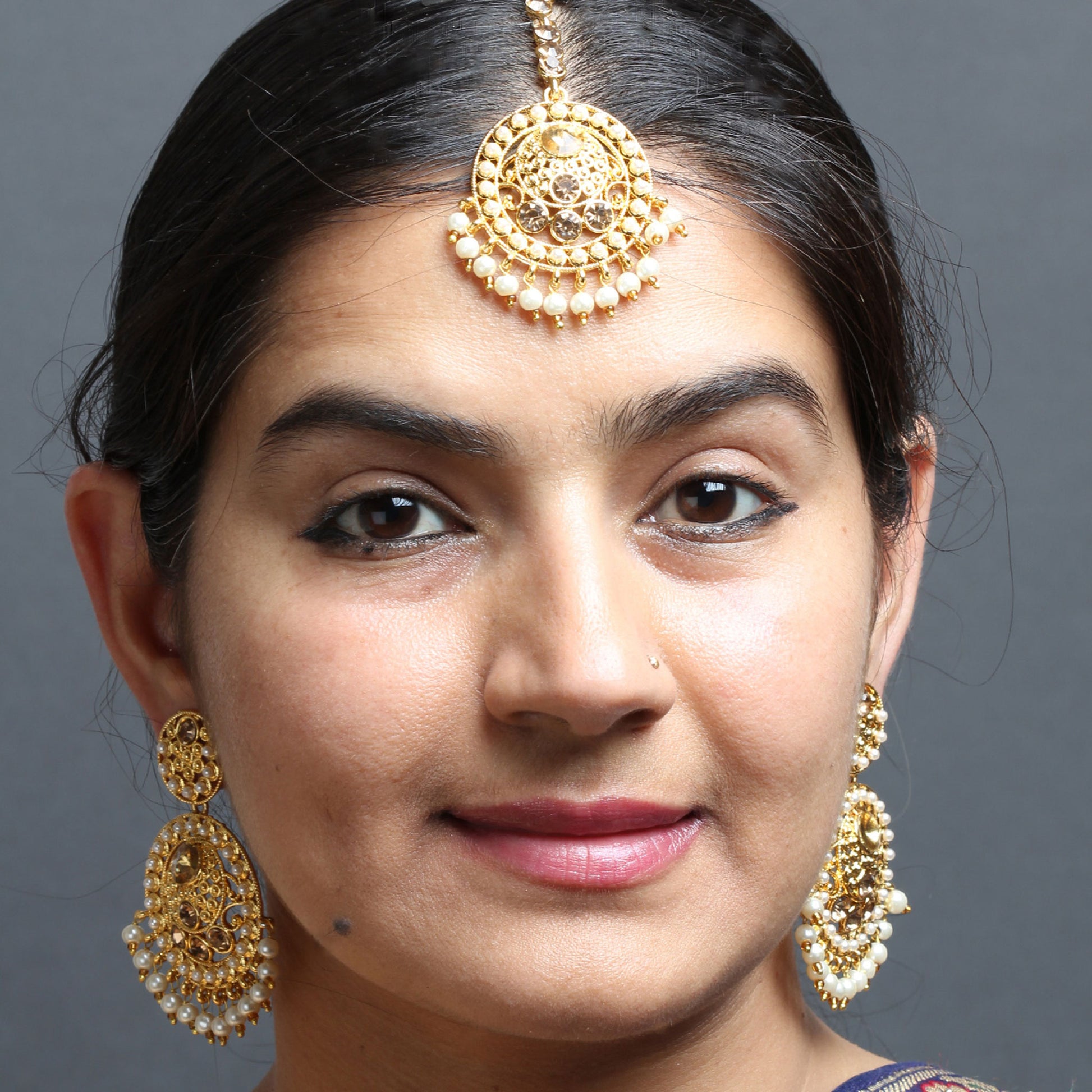 Bindhani gold-plated maang tikka and matching earrings worn on a model. The jewelry features intricate gold-toned filigree detailing with champagne-colored stones and white pearl drop accents. The maang tikka showcases a circular design with stone embellishments and a row of pearl hanging drops, while the matching earrings feature ornate circular patterns with pearl drop detailing. Close-up front view displayed against a grey background with black hair.
