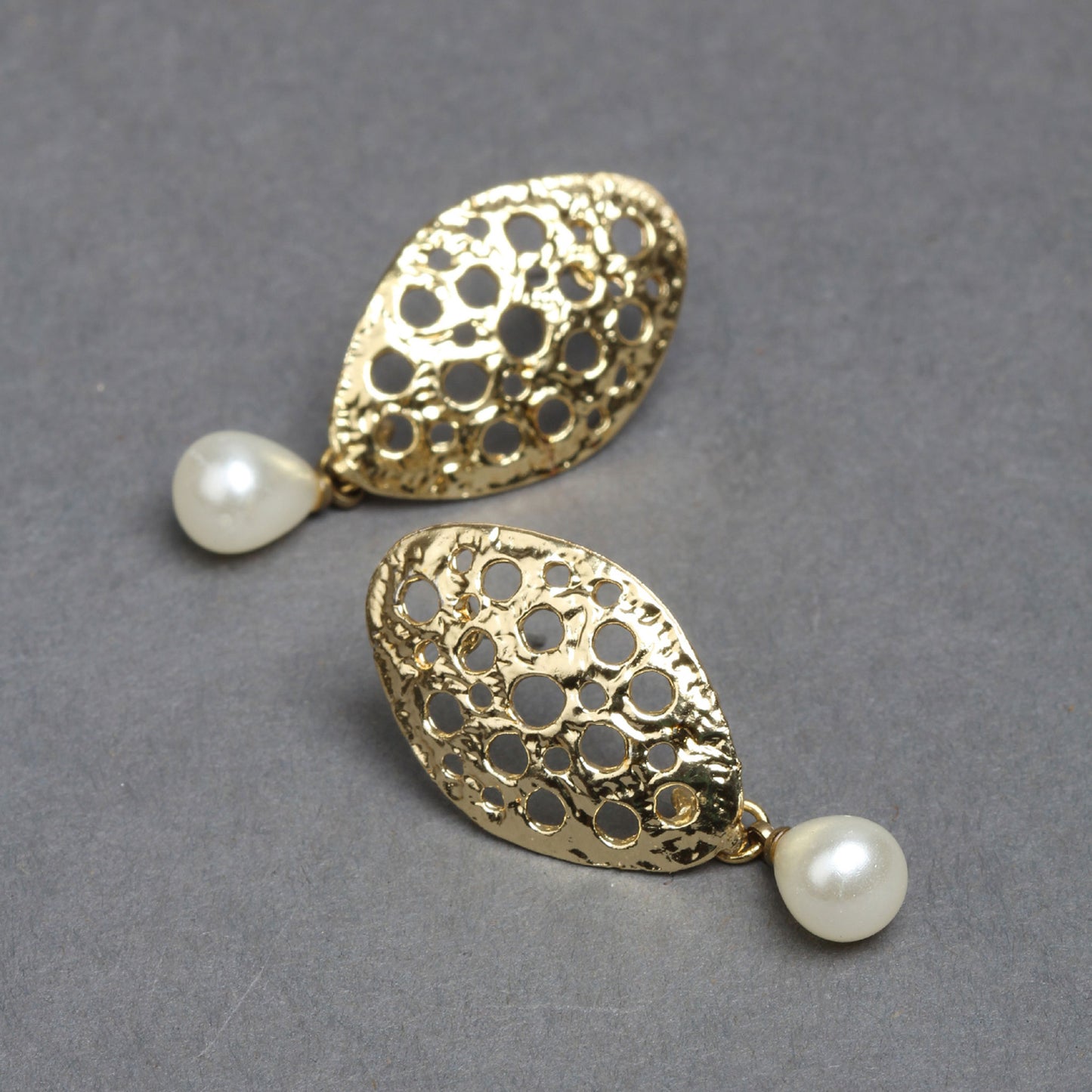 Bindhani gold-plated oval drop earrings featuring a hammered openwork design with multiple circular cutout patterns. The textured gold finish adds a contemporary look, while a single white pearl drop enhances the elegant style. Lightweight statement earrings displayed as a pair on a dark grey background.