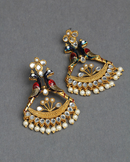 Bindhani gold-plated peacock chandelier earrings featuring two colorful enamel peacocks in blue, red, and green perched on a branch beneath a floral stud with white stones. The fan-shaped lower design is embellished with white teardrop-shaped stones, intricate gold detailing, and finished with dangling white pearl drops. Traditional handcrafted statement earrings displayed as a pair on a dark grey background.