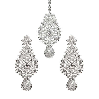 Bindhani silver-toned earrings and maang tikka set embellished with clear stones and sparkling crystal accents. The earrings feature a floral top, intricate leaf and scrollwork design with a central round stone, and a detailed teardrop-shaped stone-studded bottom drop. The matching maang tikka showcases a coordinating ornate floral design with a central round stone and a teardrop-shaped stone-studded drop. Front view displayed on a white background.