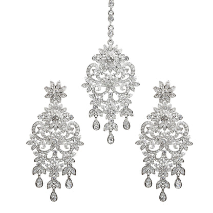 Bindhani silver-toned earrings and maang tikka set embellished with clear stones and sparkling crystal accents. The earrings feature a floral top, intricate leaf and scrollwork design with a central floral motif, and three dangling teardrop-shaped clear stone drops. The matching maang tikka showcases a coordinating floral design with detailed openwork, a central floral motif, and three dangling teardrop-shaped clear stone drops. Front view displayed on a white background.