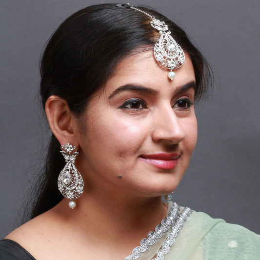 Bindhani silver-toned maang tikka and matching earrings worn on a model. The jewelry features clear stones with intricate floral and teardrop-shaped detailing along with white pearl drop accents. The maang tikka showcases an elegant teardrop design with stone-studded openwork patterns and a single pearl drop, while the matching earrings feature floral tops, teardrop-shaped stone designs, and pearl hanging drops. Close-up view displayed against a grey background