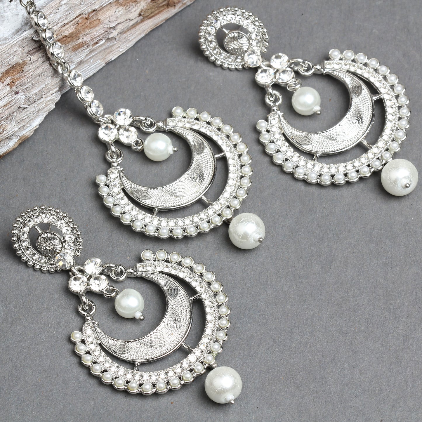 Bindhani silver-toned maang tikka and earrings set featuring white pearl detailing and clear stone embellishments. The jewelry showcases a crescent-shaped design with intricate textured detailing, pearl-studded borders, floral side motifs, and elegant white pearl hanging drops. Product displayed in close-up view on a grey background.